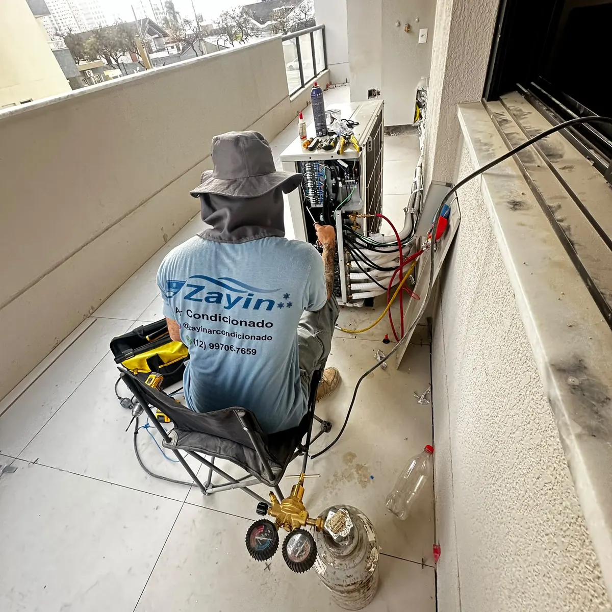 Técnico da Zayin fazendo a ligação da condensadora na sacada de um apartamento, com o manifold de pressão ao lado